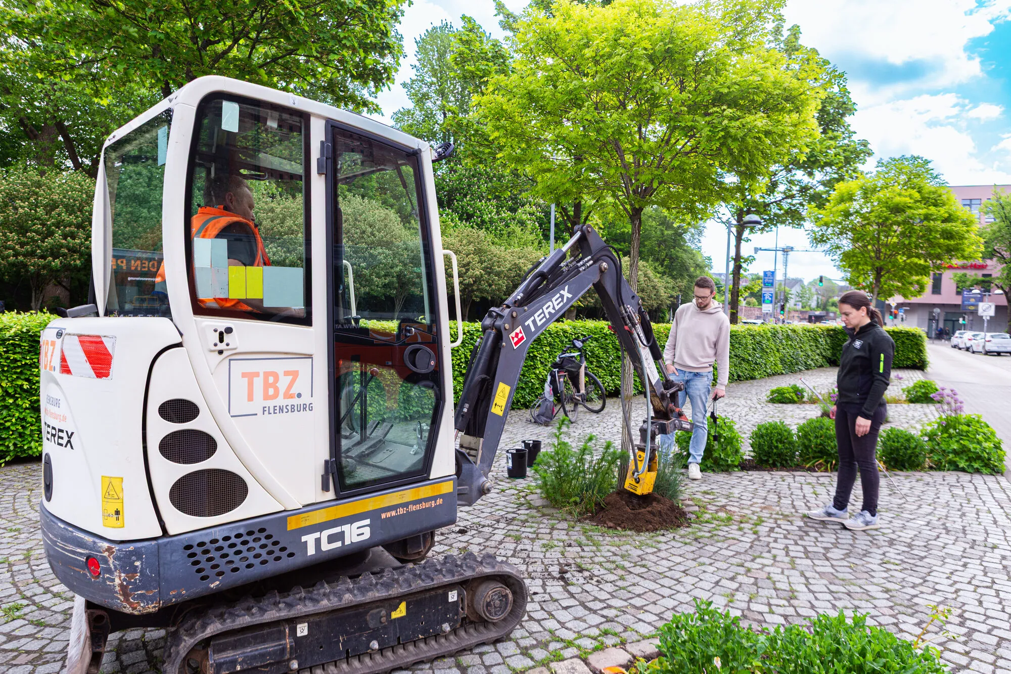 Ein Minibagger des Technischen Betriebszentrums Flensburg an einer Baumscheibe in einer Straße. Daneben stehen Cedrik Hoffmann und eine weitere Person.
