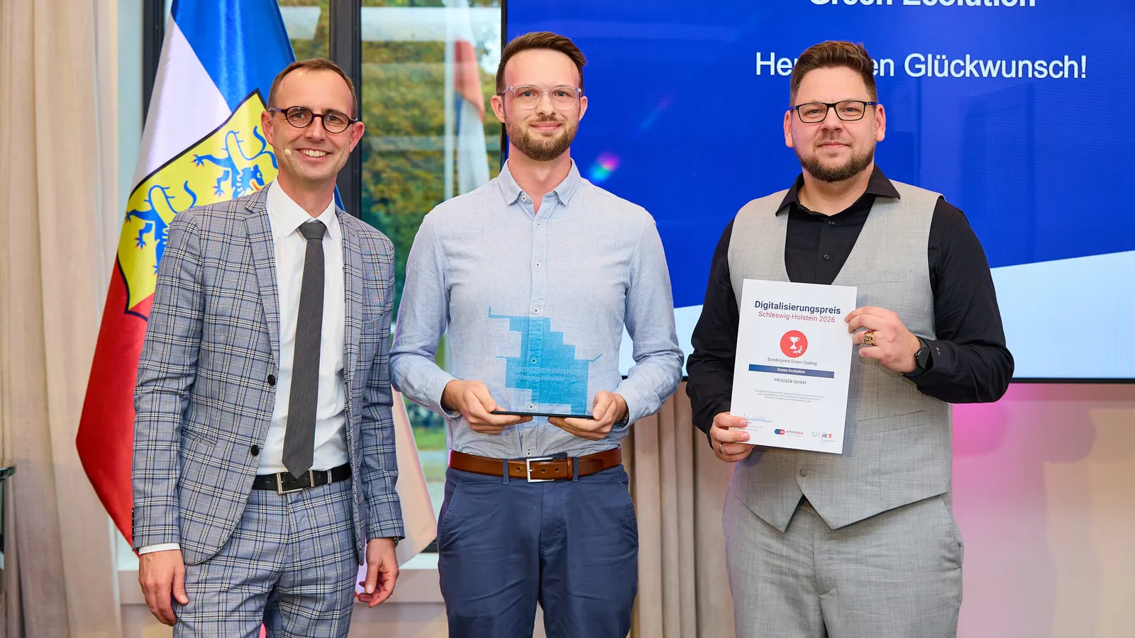Digitalisierungsminister Dirk Schrödter steht mit Cedrik Hoffmann und einem Vertreter von PROGEEK vor einer Leinwand mit der Aufschrift „Green Ecolution, Herzlichen Glückwunsch!“. Cedrik Hoffmann hält in der Mitte die gläserne Trophäe, rechts neben ihm wird die Urkunde gezeigt.
