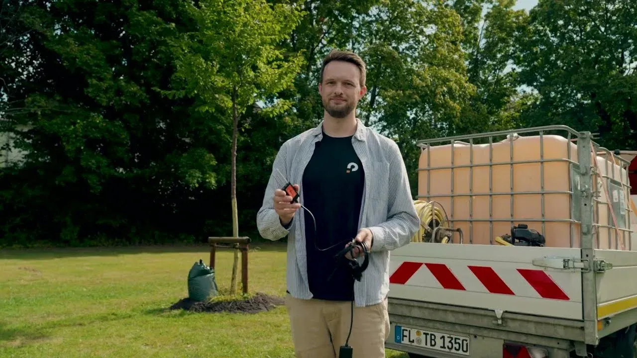 Ein Projektmitarbeiter hält einen Bodenfeuchtesensor in der Hand, im Hintergrund ein junger Straßenbaum und ein Pritschenwagen mit Wassertank
