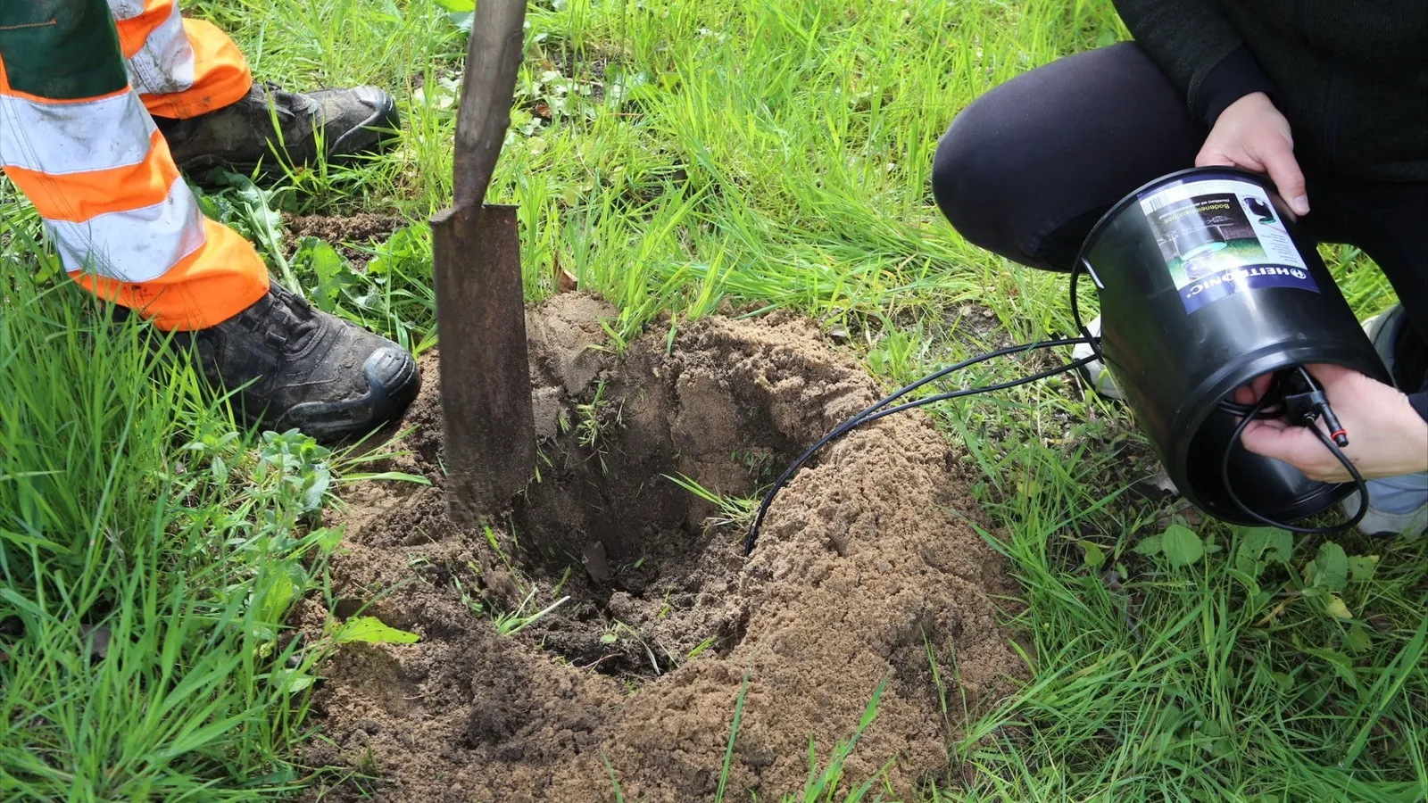 Zwei Personen setzen eine Sensoreinheit in ein ausgehobenes Loch, daneben steckt ein Spaten in der Wiese