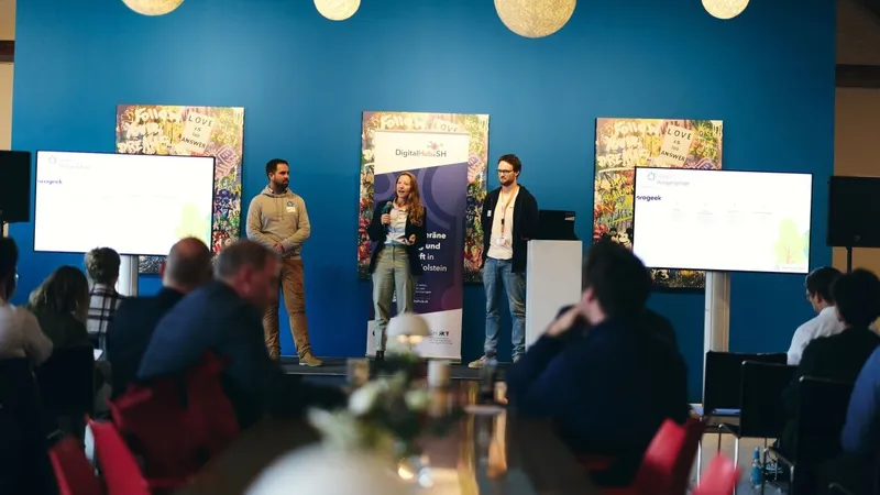 Drei Personen stellen Green Ecolution auf einer Bühne vor, rechts Cedrik Hoffmann. Zwischen zwei Bildschirmen steht ein Aufsteller des DigitalHub.SH.